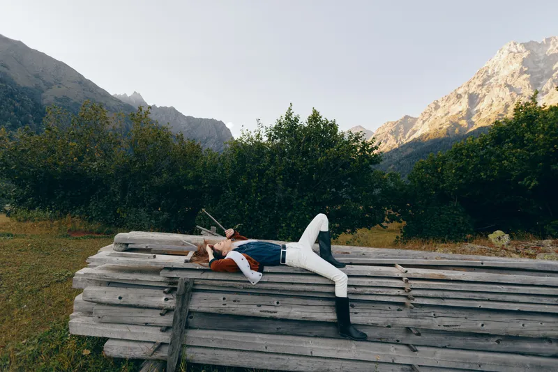 Woman resting on stacked wood beams in an orchard, surrounded by trees and mountains, outdoors relaxation scene with casual denim and boots, peaceful summer landscape.