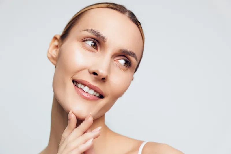 Closeup portrait of smiling young woman with natural skin looking away touching chin showing healthy glowing complexion on light background with soft expression and casual mood