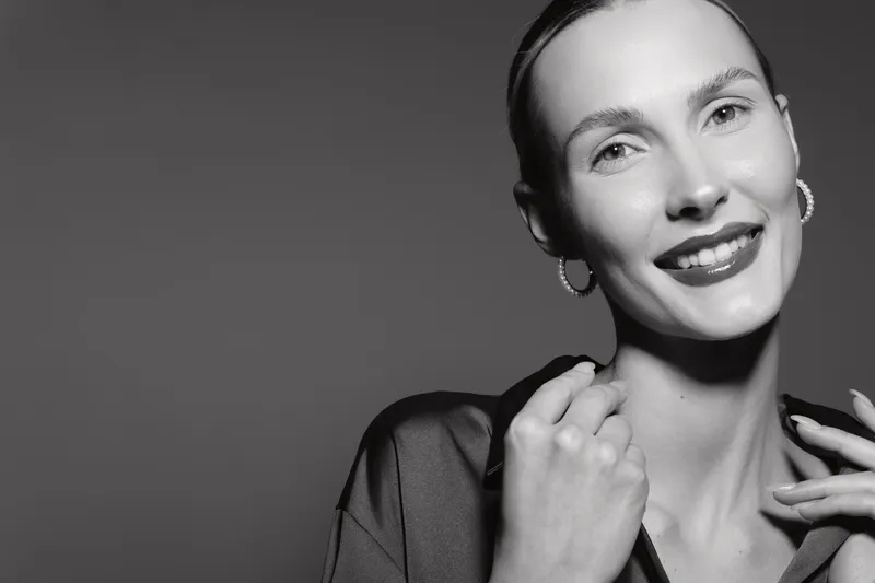 Smiling white woman with fair skin and earrings poses elegantly in black and white portrait, studio lighting.