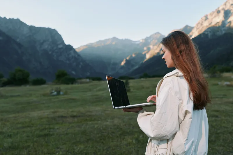 A woman with long hair stands outdoors in a valley with mountains in the background, holding a laptop and looking focused, conveying productive remote work.