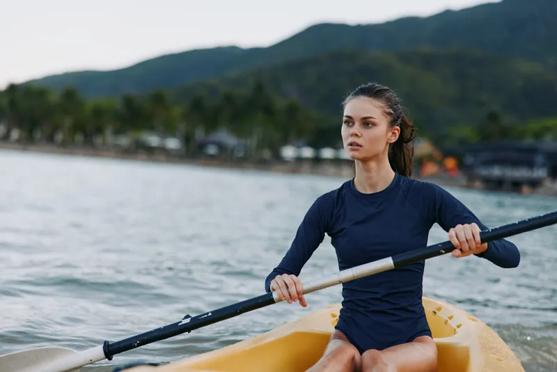 Focused kayaker paddles a bright yellow kayak on calm water, wearing dark athletic gear. Determined expression, steady stroke, sunlit backdrop, distant hills, peaceful outdoor scene.