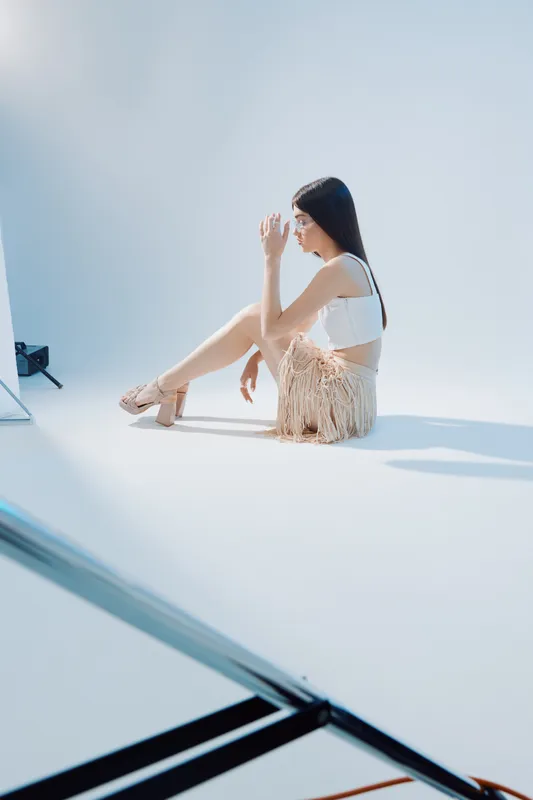 Young woman in a white top and fringed skirt sits on the floor in a bright studio. She poses thoughtfully with long dark hair and elegant sandals. Concept of beauty fashion.