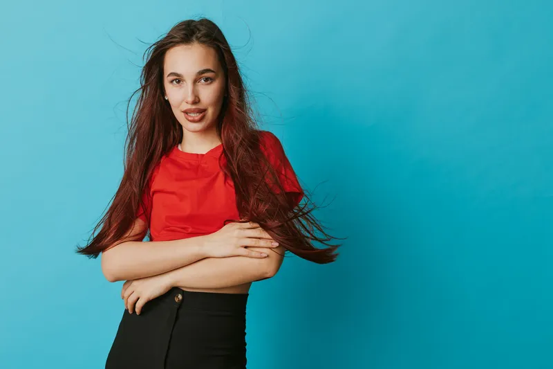 Young woman with long hair wearing red shirt and black skirt poses with arms crossed against blue background. Casual style, confident expression and natural look in studio portrait.