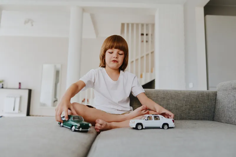 Young boy playing with toy cars on a couch in a bright living room, showcasing creativity and imagination while having fun.