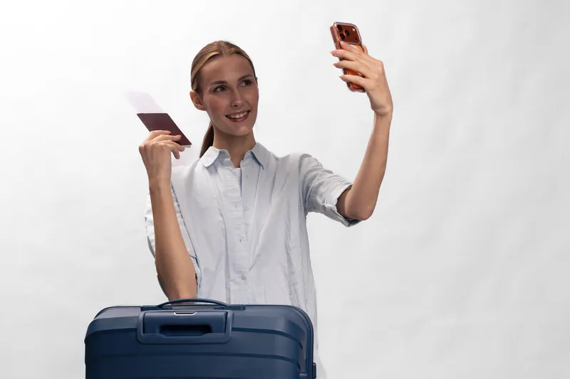 Young woman with suitcase holds passport and phone taking selfie on white background. Concept of travel, vacation, tourism, and happy female traveler before trip.