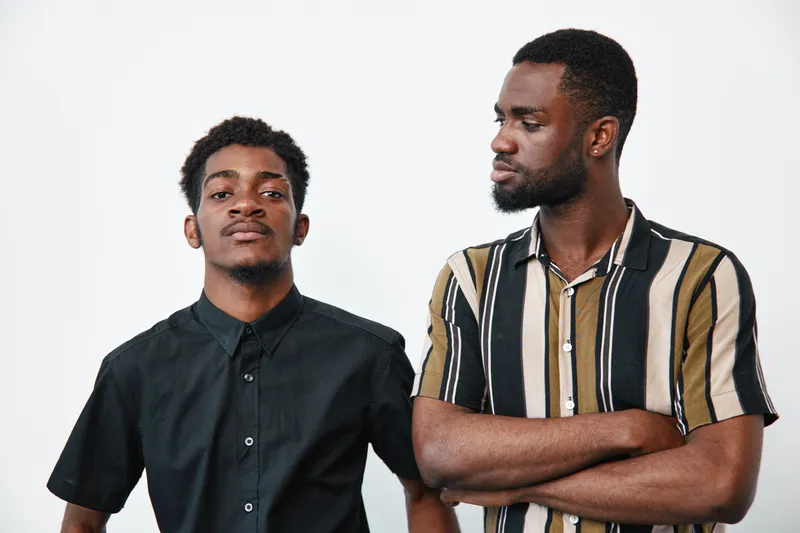 Two African American men with dark skin standing against white background, one wearing black shirt and the other striped shirt with arms crossed, looking confident and serious.