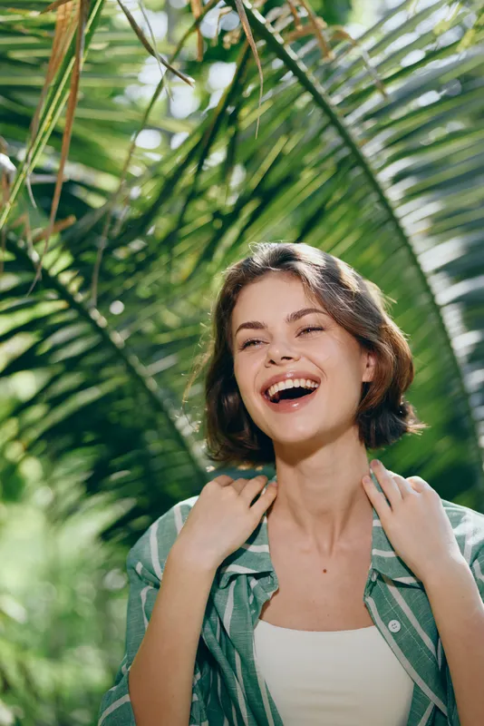 Vibrant portrait of a smiling woman in a sunlit tropical setting, framed by lush greenery and palm leaves.