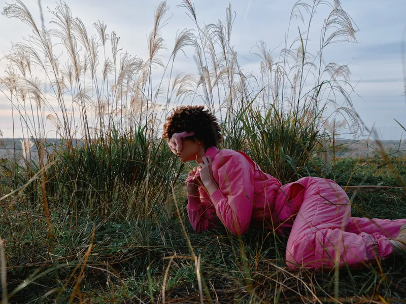 Woman in a pink outfit lying in tall grass, outdoor nature scene, relaxed pose and tranquil mood, soft light and casual lifestyle in a green field.