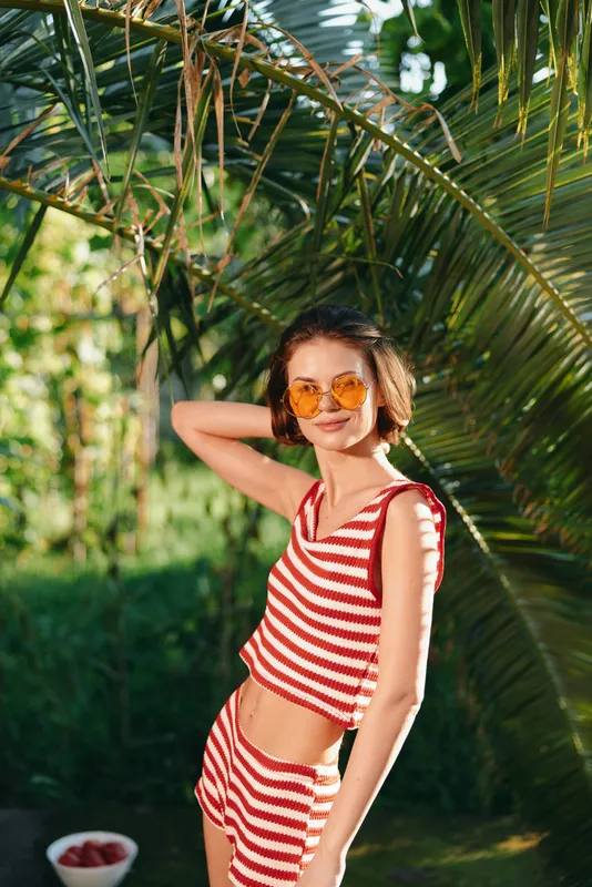 A cheerful woman wearing a red and white striped swimsuit stands in a sunny tropical garden, posing with hands on hips, radiating confidence, joy, and carefree summer energy in a vivid outdoor scene.