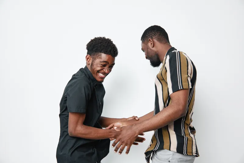 Two African American men with dark skin laughing and shaking hands in casual clothes on white background. Friendship, happiness and connection between young black males.