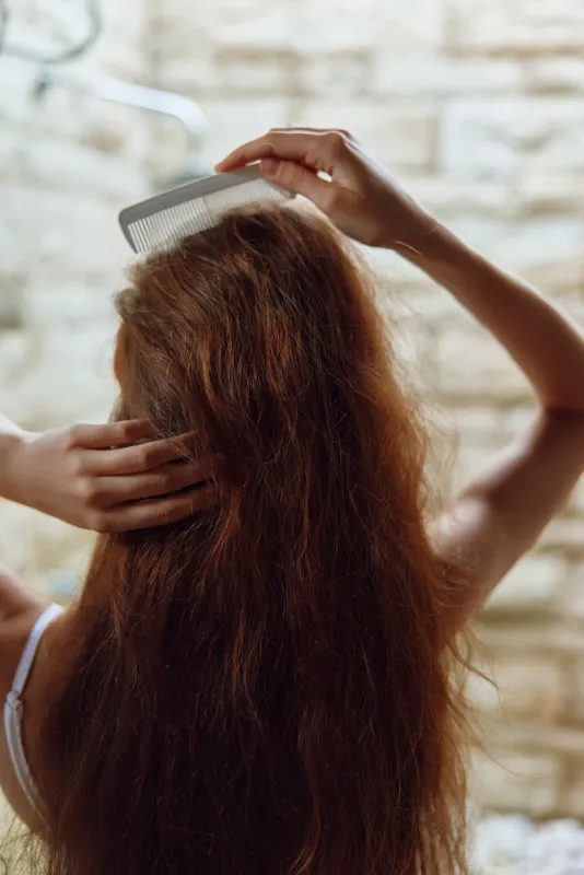 woman combing hair with white comb, long brown hair, natural light, casual indoor setting, textured brick wall background, hair care routine