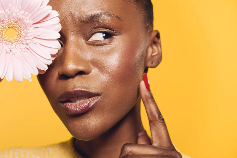 Close-up of thoughtful dark skinned African woman touching her cheek with finger near pink flower on yellow background. Concept of skincare, beauty, and natural cosmetics for black skin.