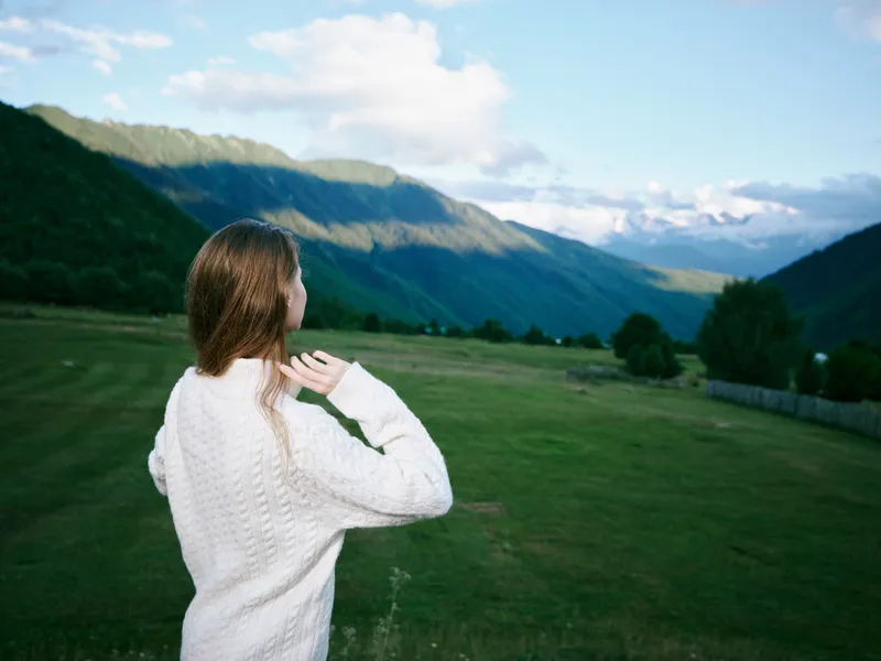 Landscape scene of a woman in a white sweater facing distant mountains and a green valley under a blue sky, conveying exploration, calm, nature, and outdoor freedom.