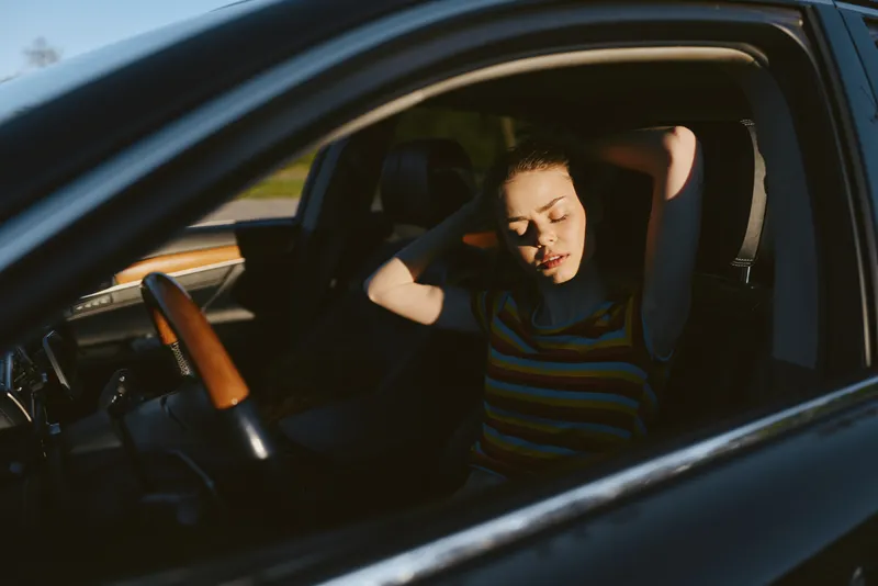 Woman relaxes inside a car with sunlight on her face, striped shirt, casual mood, retro film color palette, warm interior textures, lifestyle portrait with calm atmosphere.