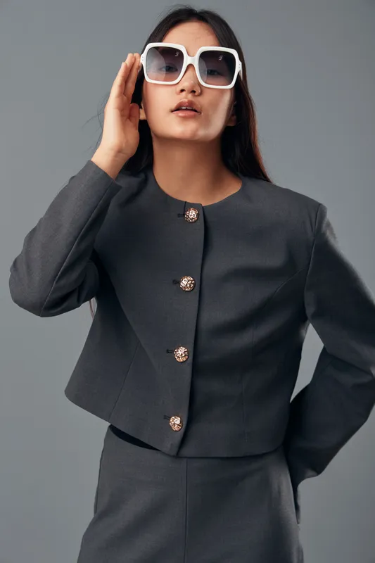 Woman wearing sunglasses with modern dark gray suit posing confidently against a plain gray background. Fashion model with straight hair showcasing stylish eyewear and tailored outfit.