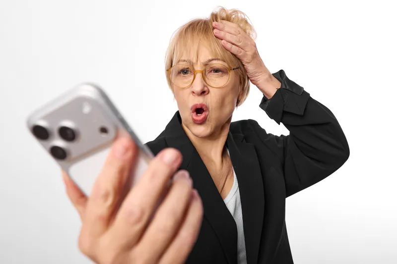 Woman elderly shocked with glasses holding a smartphone close to camera, surprised mouth open, hand on head, studio white background, technology portrait