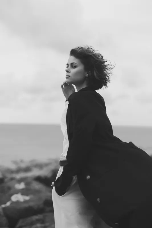 Woman on a windy coastline, flowing coat, thoughtful gaze, sea breeze in her hair, captured in grayscale to emphasize mood, solitude and the drama of the shore, weathered rocks and open horizon