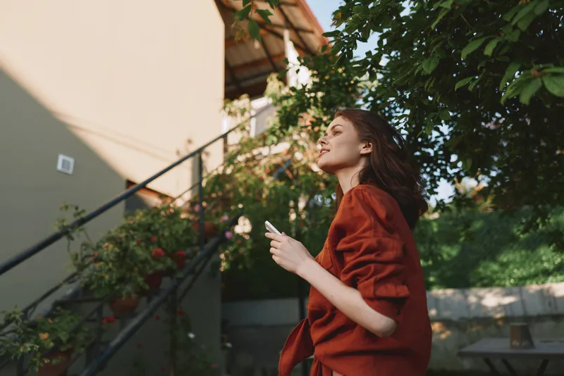 A woman in a red coat stands outdoors, looking up with a phone in hand. A calm, sunny day frames a casual street style and reflective mood