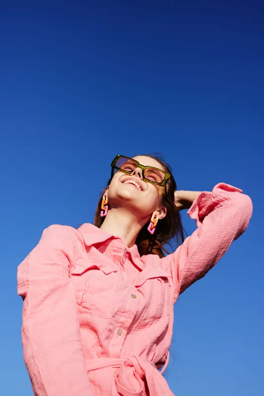 Joyful young woman in stylish pink outfit against clear blue sky, showcasing summer fashion trends and vibrant colors for a fresh lifestyle concept