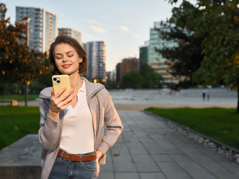 A cheerful young woman stands outdoors in an urban park, smiling at her smartphone as sunlight highlights her casual attire and confident posture, creating a bright, contemporary vibe for lifestyle,
