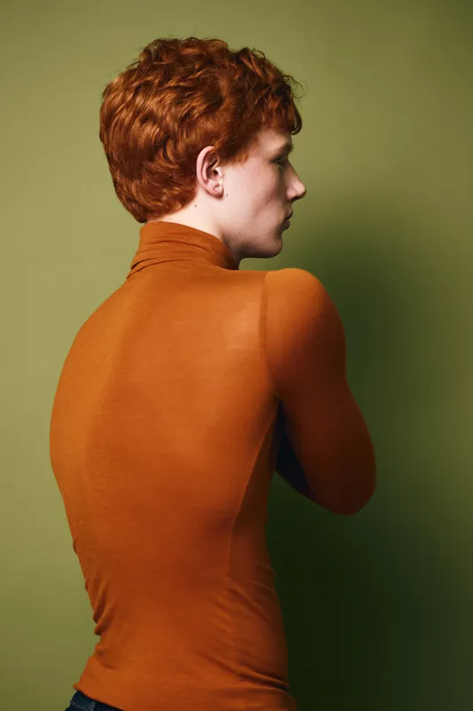 Portrait of young redhead person with curly hair wearing orange turtleneck, standing against olive green background, viewed from side with crossed arms and serious expression.