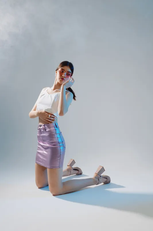 Young woman in a white top and shiny pink skirt poses on her knees in a studio with soft lighting. Concept of beauty, fashion, and modern style with elegant makeup and accessories.