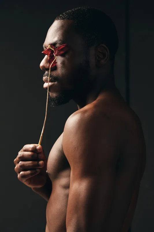 Creative conceptual portrait of African man with dark skin holding dried flower in front of eyes, artistic visual style with dramatic lighting and minimalistic background in studio.