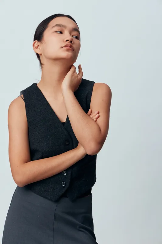 Young woman wearing a black sleeveless vest and skirt posing against a plain background with a thoughtful expression and hand touching her neck. Studio portrait of confident fashion model.