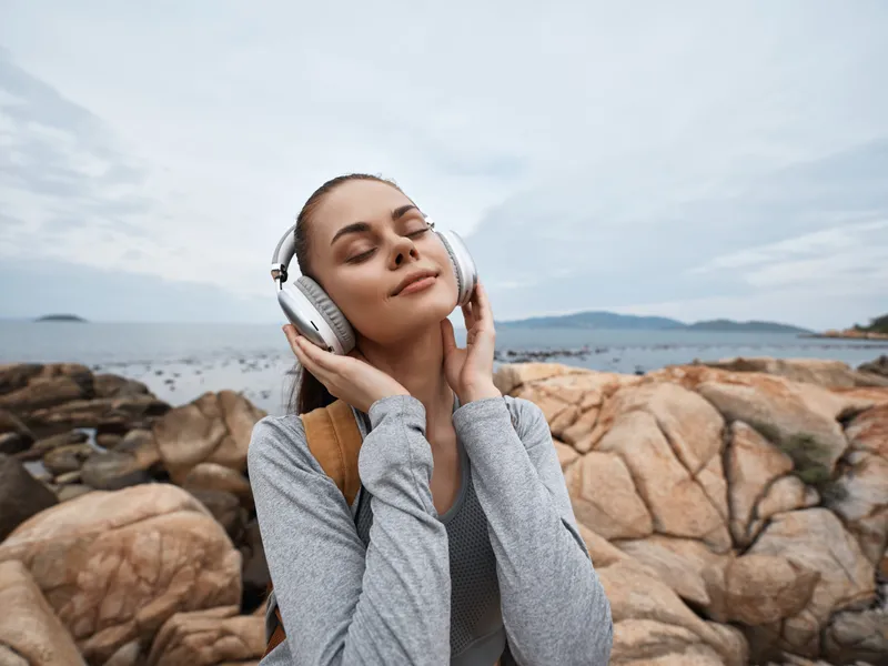 A young woman with fair skin enjoys music by the seaside, wearing white headphones and a casual gray outfit, evoking feelings of relaxation and connection with nature