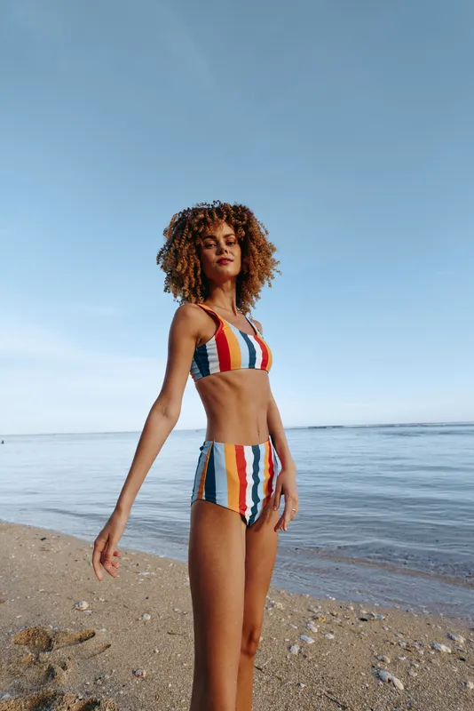 A confident young woman in a colorful striped bikini stands on a sandy beach near calm sea water.