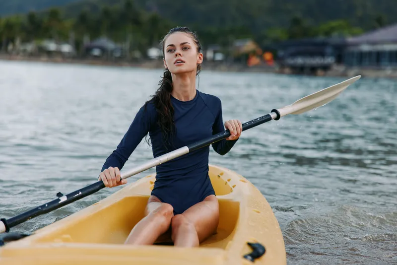 Young woman paddling a yellow kayak on calm water, focused expression, long hair tied back, navy long sleeve, capturing outdoor adventure and athleticism.