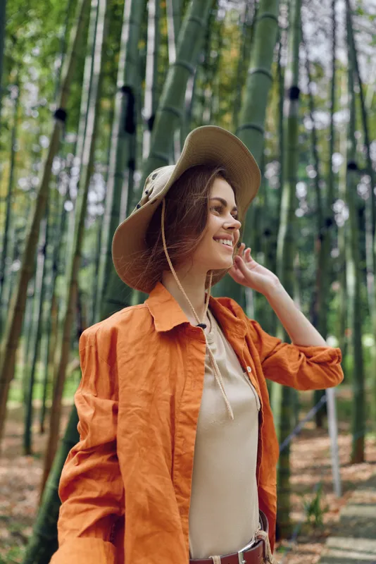 Woman wearing hat and orange jacket smiles in bamboo forest portrait, enjoying nature and travel moment among tall green stalks, casual outfit and relaxed outdoor mood