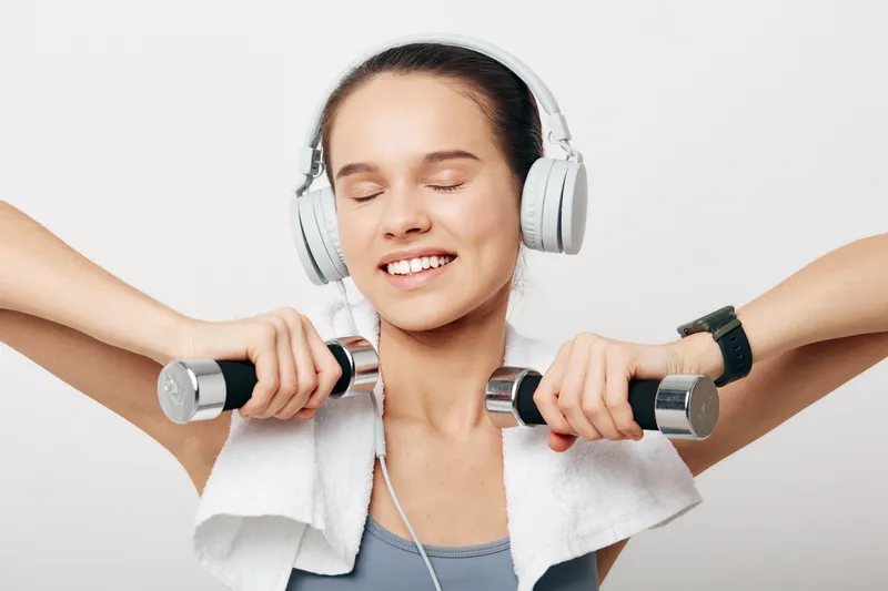 Fitness woman wearing headphones lifts dumbbells with a towel draped over shoulders, smiling through a focused workout in a bright studio, showcasing strength, determination and energy.