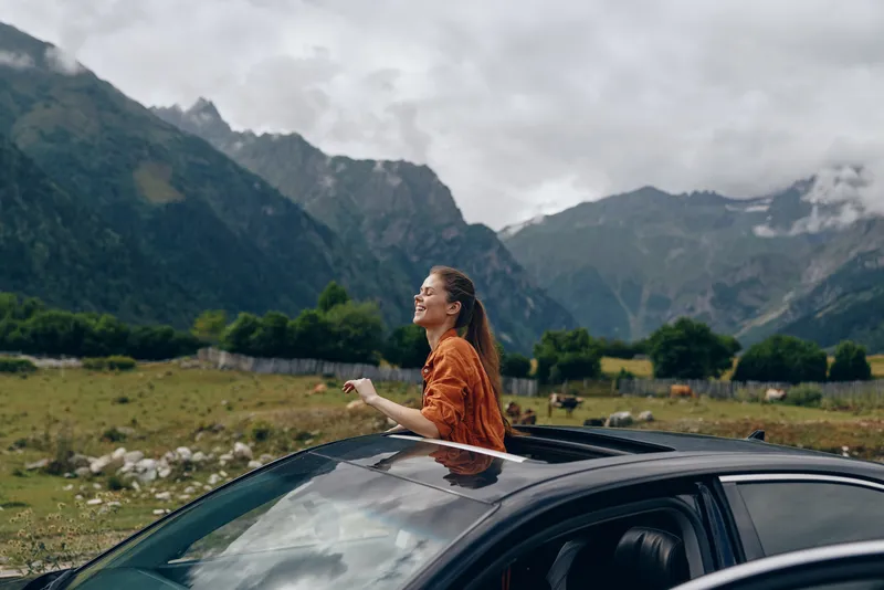 A woman leans from a car sunroof in a wide open landscape, mountains in the background, enjoying fresh air and scenic countryside.