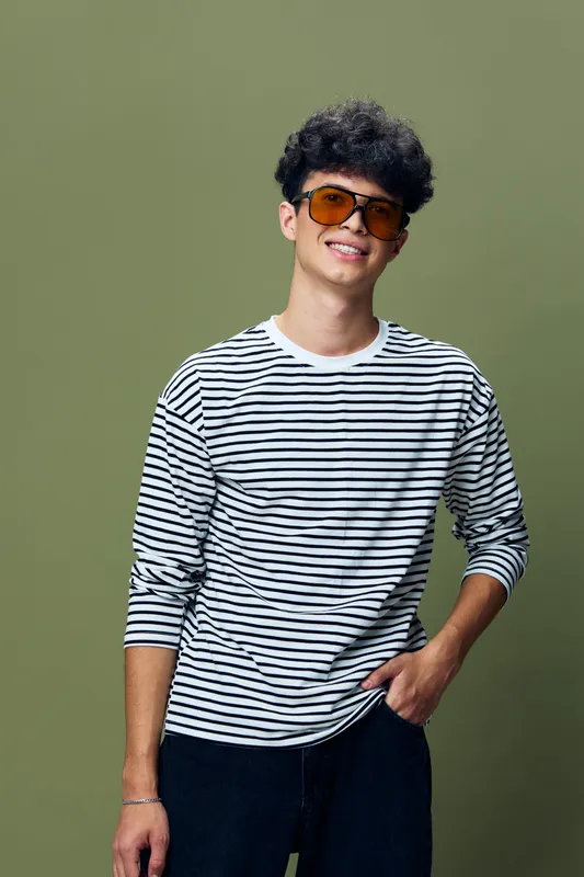 Smiling young man wearing striped shirt and orange sunglasses stands against olive background with relaxed posture, casual style, and joyful expression.