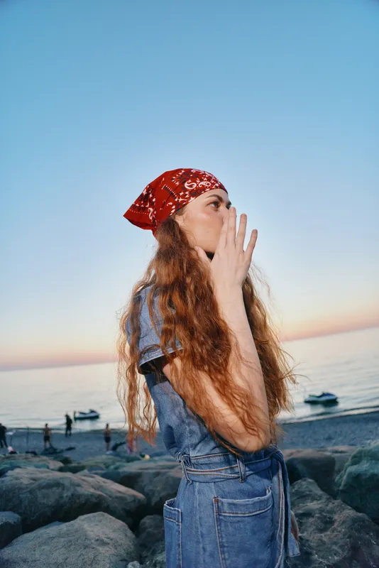 young woman wearing red bandana and denim outfit standing on rocky beach during sunset with calm sea background