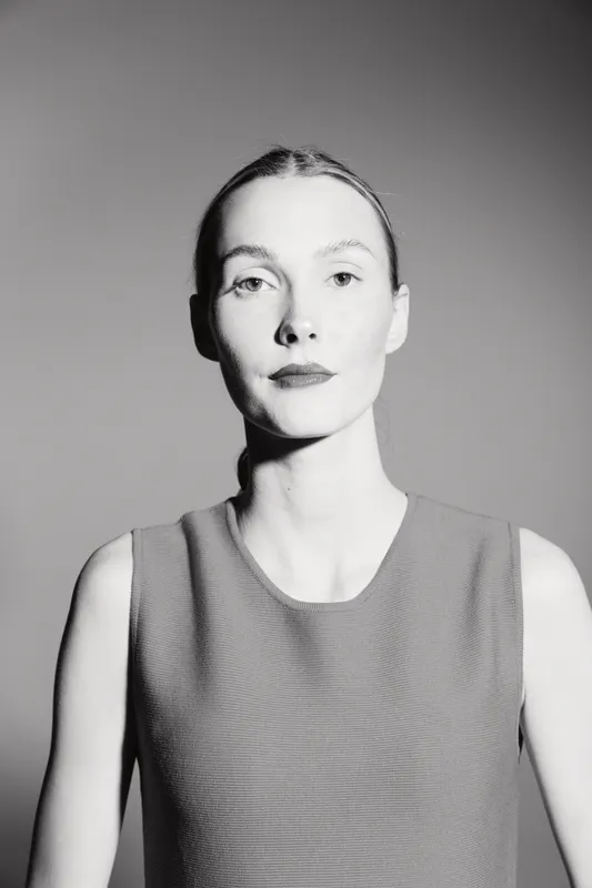 Portrait of a pale white woman with blonde hair in a sleeveless top looking confidently at the camera. Minimalist black and white studio lighting highlights her facial features.