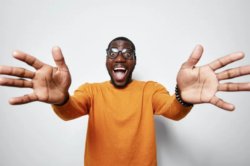 African American man with dark skin wearing glasses and orange sweater smiling widely with open arms on white background for happiness and excitement concept.