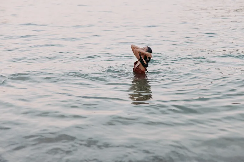 Person in calm water lifts a hat above the surface, creating a serene moment of playful solitude as light dances on ripples around them.