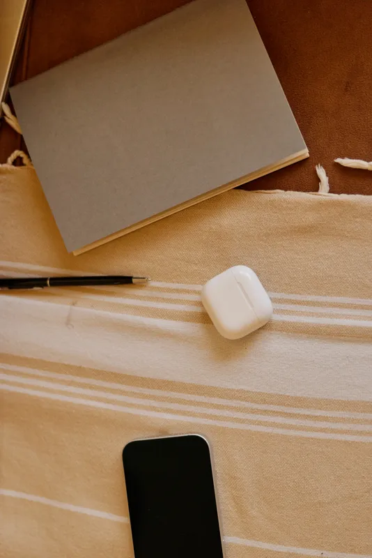 Cozy home lifestyle scene with a notebook, pen, wireless earbuds case and smartphone resting on a soft striped blanket beside a bed creating a calm desk setup