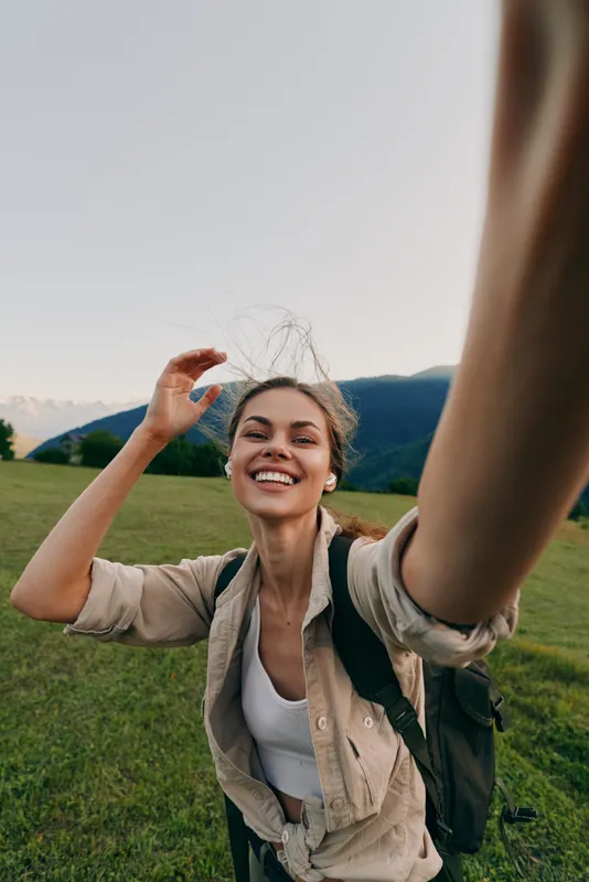 Young woman takes a bright selfie in a green meadow, smiling as wind lifts hair and a backpack rests on her shoulders.
