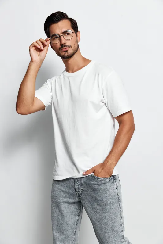 Stylish young man wearing a white t-shirt and glasses poses confidently against a solid light background, showcasing a trendy urban fashion style.