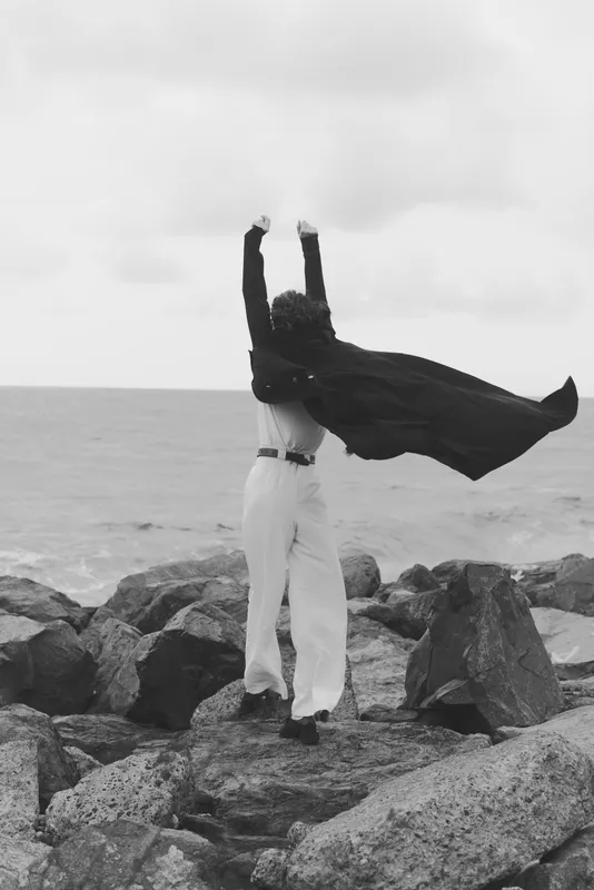 Silhouette of a figure in a cape on a rocky shore, arms raised toward the sky in an outdoor dramatic moment, conveying strength, freedom and resilience against the sea breeze