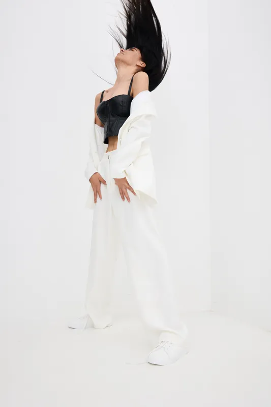 young woman in motion with long black hair, wearing white wide-leg pants and black crop top, standing against white minimalist background in dynamic pose