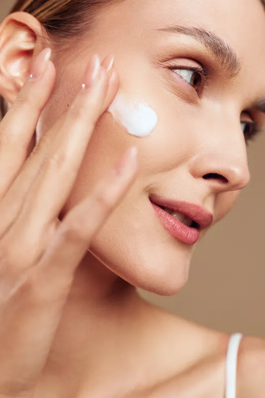 Young woman with clear skin applies moisturizing cream on her cheek using fingers in a close-up beauty skincare routine against a neutral background.