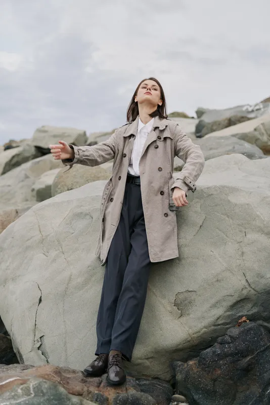 A confident woman in a long coat stands on large rocks outdoors under a cloudy sky. The scene conveys strength, freedom, and calm in a natural rocky environment.
