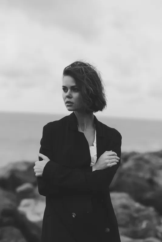 Woman in a black blazer stands with folded arms on a rugged coast, moody monochrome image conveying resilience, solitude, and coastal atmosphere, wind and sea spray accentuate texture and mood