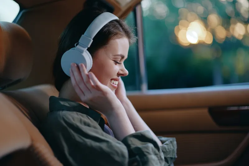 Young woman enjoying music in a car, wearing headphones, smiling joyfully, against a blurred nature background in warm tones, perfect for music enjoyment themes.