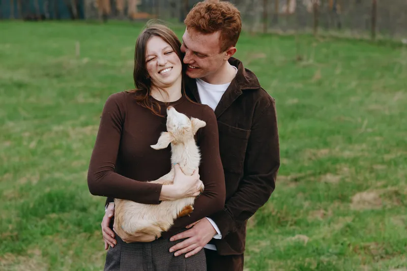 A smiling couple holds a goat kid in a grassy meadow, sharing a warm outdoor moment. A lifestyle scene that celebrates affection, companionship, and simple farm life in daylight.