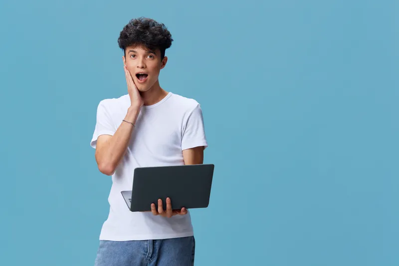 surprised young man holding laptop with open mouth and hand on cheek, wearing white t-shirt and jeans against blue background, casual technology concept